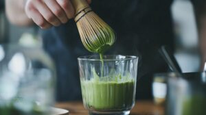 A close-up of a traditional bamboo whisk (chasen) lifting freshly whisked matcha from a steaming glass