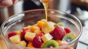 Close-up of honey being poured over a bowl of mixed fruits, including strawberries, kiwi, and pineapple
