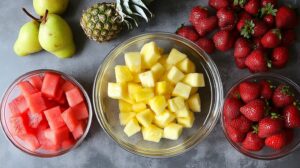 Bowls of chopped pineapple, watermelon, and strawberries surrounded by whole pears, pineapple, and strawberries