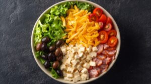 A bowl of salad with lettuce, shredded cheddar cheese, cherry tomatoes, olives, feta cheese, and sliced deli meat arranged in sections