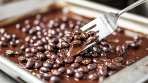 Close-up of coffee beans being coated in melted chocolate on a tray, with a fork lifting some beans