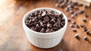 A bowl of chocolate chips on a wooden surface, with scattered chips around