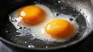 Two sunny-side-up eggs cooking in a black frying pan with bubbling egg whites and vibrant yellow yolks