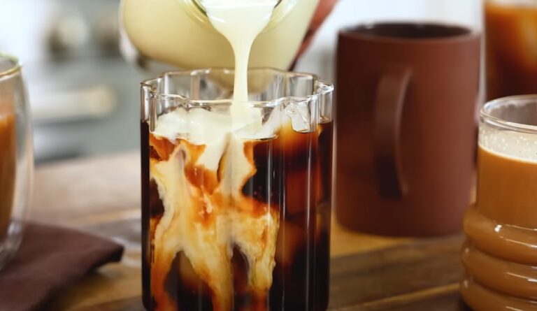 A milk creamer is gently added to a glass of iced coffee