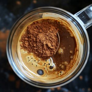 Overhead view of a clear glass mug with coffee and a heaping spoon of cocoa powder, partially mixed, highlighting the start of a flavorful coffee enhancement