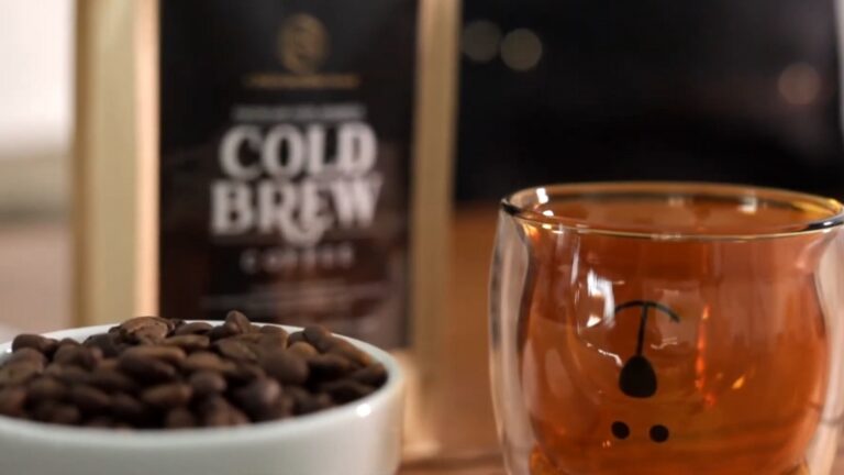 A bowl of coffee beans, a cup of cold brew coffee, and a cold brew coffee bag in the background
