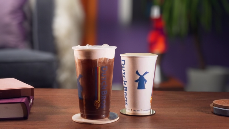 Two Dutch Bros drinks, one iced and one hot, sit on a wooden table in a cozy indoor setting