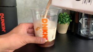 Milk is being poured into a Dunkin' cup filled with ice and caramel syrup while someone holds the cup