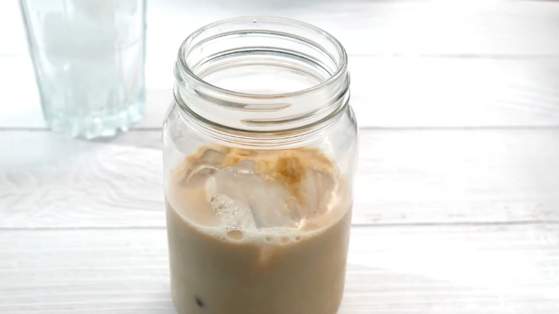 A glass mason jar filled with iced coffee and ice cubes sits on a white wooden surface
