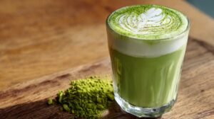 Glass of iced matcha latte with latte art on top, sitting on a wooden surface next to a small pile of matcha powder