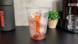 A clear Dunkin' cup filled with ice and caramel syrup sits on a black countertop next to a coffee maker