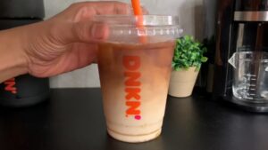 A hand holds a Dunkin’ iced coffee with visible layers of milk, coffee, and syrup, topped with an orange straw
