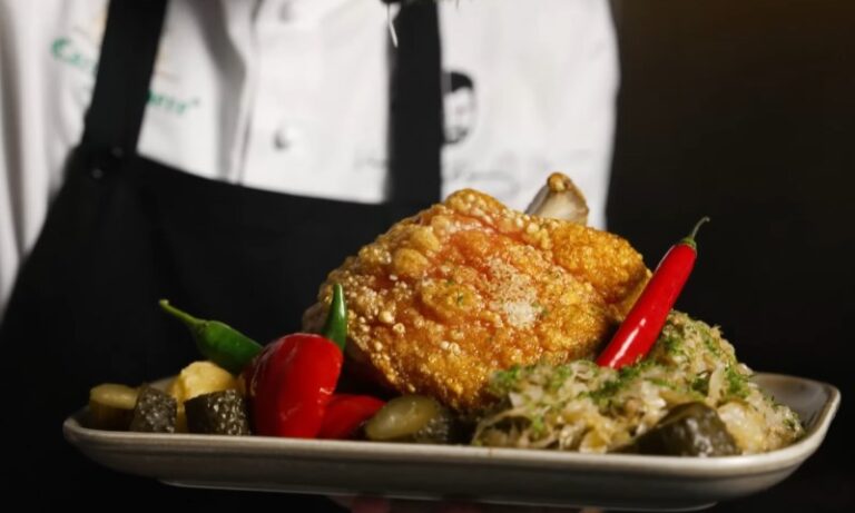 A chef in a white jacket and black apron holds a platter with crispy pork, vibrant pickled vegetables, and sauerkraut