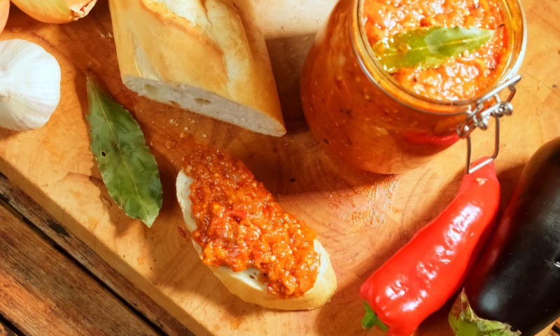 A rustic cutting board features a slice of baguette with red pepper spread, a jar of the same spread